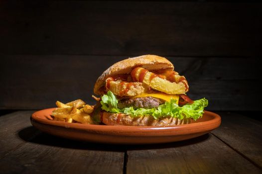 Premium quality burger with onion rings, cheese and barbecue sauce, served with french fries on a rustic plate. High quality photography.