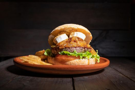 Premium quality Beef burger with goat cheese, caramelized onions in a rustic bun with french fries on the side. Yummy Burger served on a rustic plate on wooden table. High quality photography