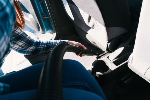 Detail of a young girl in a blue shirt and jeans vacuuming the car seat. Photographed in natural light from outside the car.