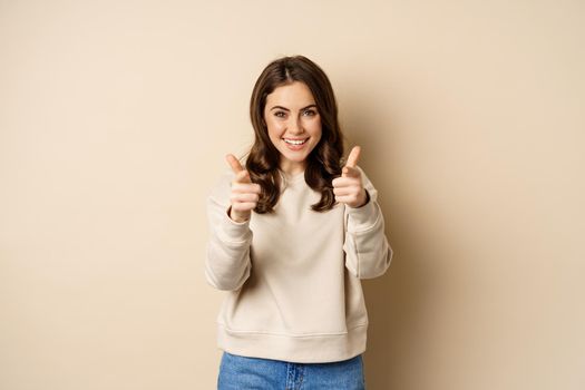 Smiling happy woman choosing you, pointing fingers at camera and laughing, inviting people, standing over beige background.