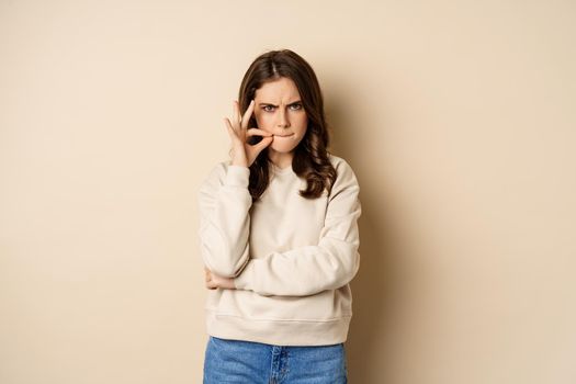 Angry woman shows mouth zipper, seal lips gesture, standing over beige background, taboo sign.