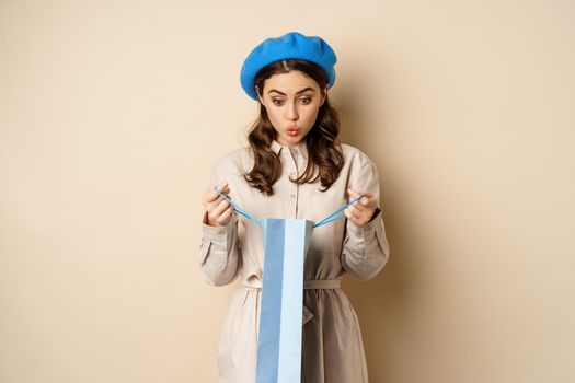 Image of girl looking surprised after opening gift bag, looking at present amazed, standing in stylish outfit over beige background.