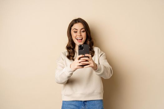 Happy woman recording video, shooting photo on smartphone camera and smiling, standing over beige background.