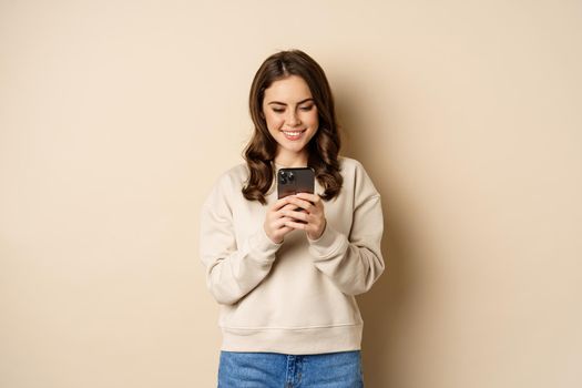 People and cellular technology. Beautiful stylish woman using mobile phone, smartphone app, smiling and looking at screen, standing over beige background.