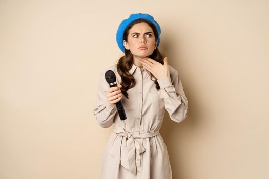 Stylish singer, woman with microphone touching throat, feeling ill, standing over beige background. Copy space