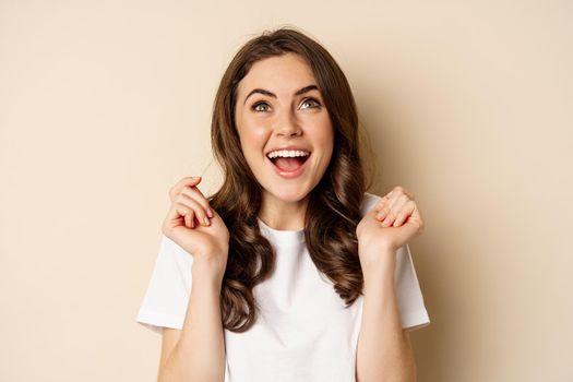 Close up portrait of enthusiastic young woman rejoicing, shouting with joy and satisfaction, celebrating victory, winning and triumphing, standing over beige background.