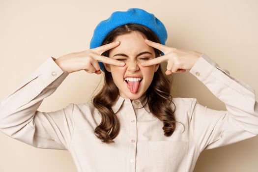 Close up portrait of carefree happy woman, showing peace v-sign and laughing, having fun, standing against beige background.