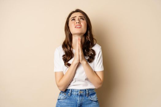 Woman pleading, begging and looking up desperate, need help, standing over beige background.