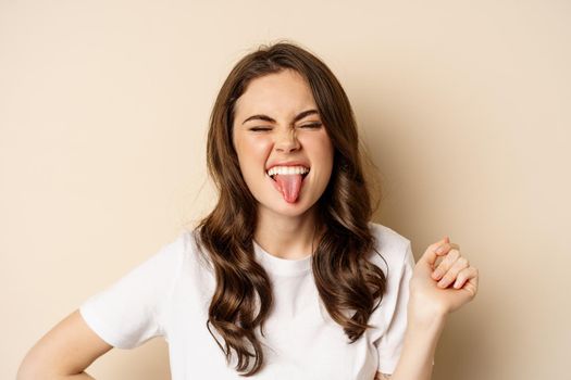 Close up of happy, carefree beautiful woman posing silly, showing tongue, having fun, standing in casual white t-shirt against beige background.