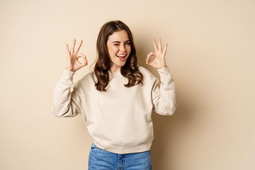 Very good, nice work. Cheerful young woman agrees, shows okay, ok praise, recommending smth, standing over beige background.