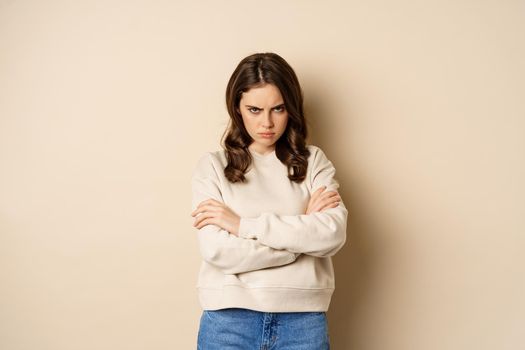 Angry girlfriend sulking, cross arms on chest and look offended, staring moody at camera, standing over beige background.