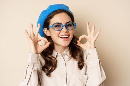 Image of happy smiling woman in stylish sunglasses, showing okay signs in approval, recommending eyewear store, beige background.