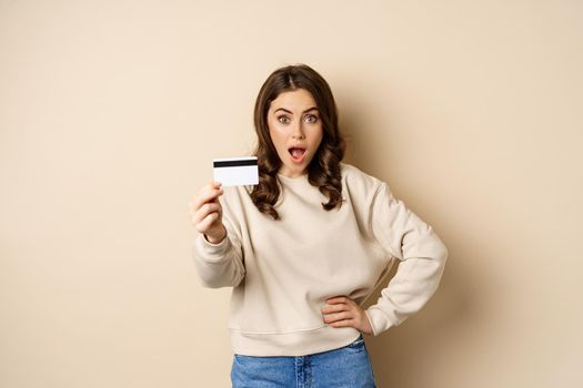 Surprised woman showing credit card, gasping impressed, impression of discounts or shopping, standing over beige background.