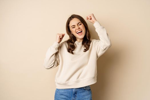 Enthusiastic brunette woman laughing and smiling, cheering, shaking hands and celebrating, triumphing, standing over beige background.