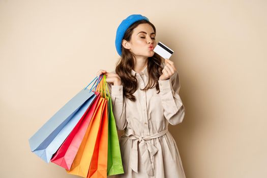 Portrait of cute happy girl kissing her credit card after buying gifts, holding shopping bags, standing over beige background. Copy space