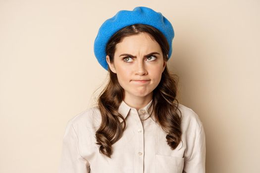 Close up portrait of young beautiful woman sulking, looking angry and disappointed, grimacing displeased, standing over beige background.