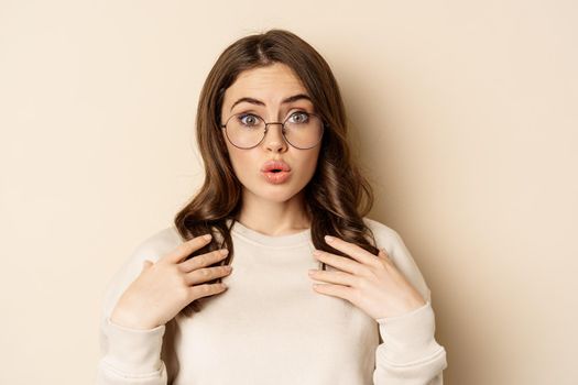 Close up portrait of beautiful girl in glasses, looking amazed and surprised, gasping excited, standing over beige background.