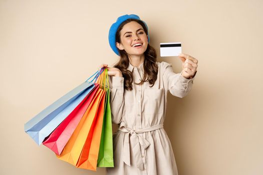 Portrat of trendy feminine girl posing with shopping bags from store and credit card, paying contactless, buying with discount on sale, beige background.