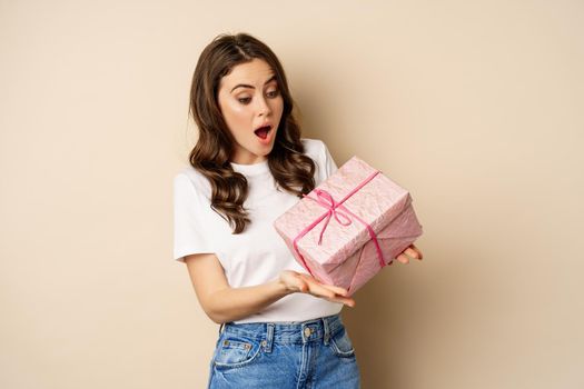 Celebration and holidays concept. Happy young woman holding gift wrapped in pink box, receive present, looking amazed and surprised, standing over beige background.