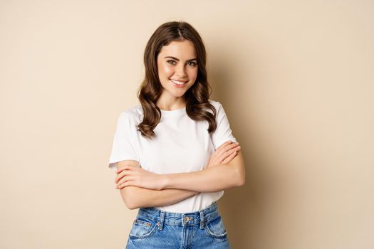 Portrait of beautiful young woman in white t-shirt, smiling and looking happy, posing against beige background.