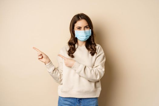 Health and covid-19 concept. Cute brunette woman in medical face mask, pointing fingers left at logo, banner, showing copy space, standing over beige background.