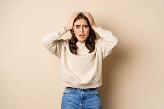 Frustrated and troubled woman facing disaster, holding hands on head in panic, looking anxious, standing over beige background.