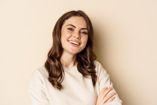 Close up portrait of young caucasian woman with dark hair, smiling white teeth, laughing, posing carefree against beige background.