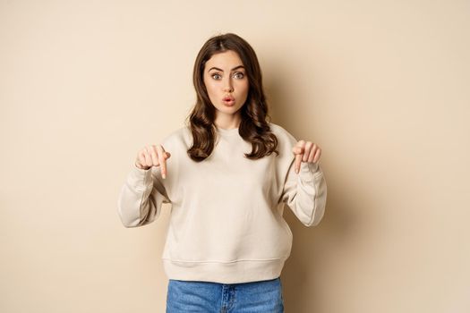 Enthusiastic attractive girl pointing fingers down, showing advertisement, link or promo below, standing over beige background.