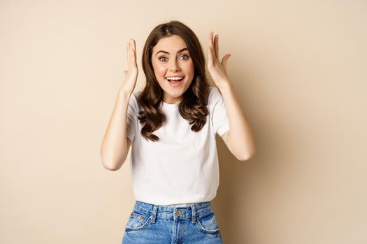 Enthusiastic adn surprised woman winning, dancing and celebrating, standing in tshirt over beige background.