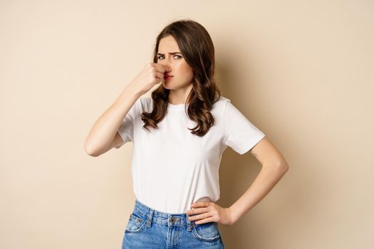 Woman shut nose from disgusting smell, standing over beige background.