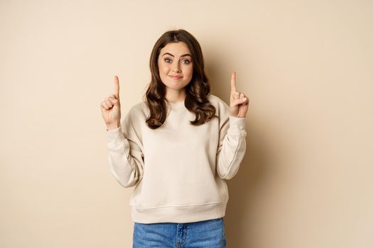 Smiling young woman looking indecisive, shrugging and smirking while pointing fingers up, standing over beige background.