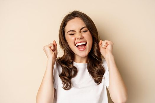 Close up portrait of enthusiastic young woman rejoicing, shouting with joy and satisfaction, celebrating victory, winning and triumphing, standing over beige background.