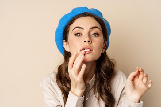 Close up portrait of young beatufiul woman looking in mirror and fixing her makeup, put on lipstick, standing over beige background.