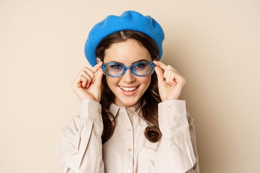 Close up portrait of stylish woman posing in sunglasses, laughing and smiling, wearing trendy outfit, standing over beige background.