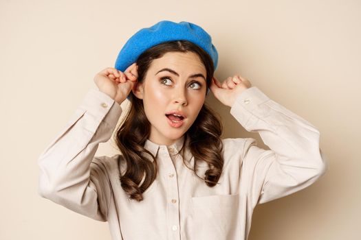 Stylish modern girl put on trendy hat on head and smiling, going out, posing against beige background.