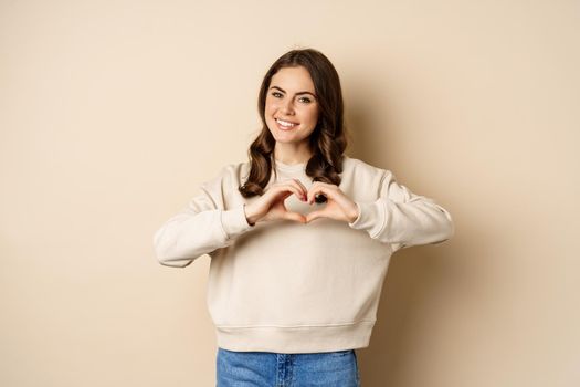 Beautiful, romantic brunette woman showing heart, love sign, care for you, smiling at camera, standing over beige background.