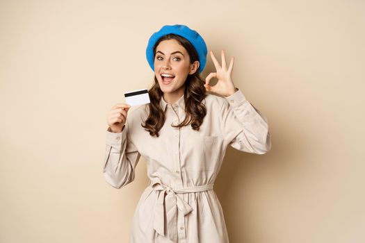 Concept of shopping and sale. Happy young woman in stylish clothes, showing credit card store discount and okay, satisfied, like and approve smth, standing over beige background.