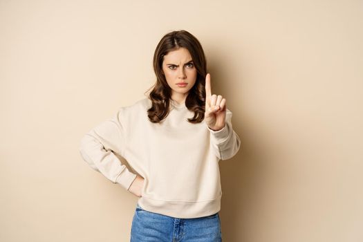 Serious woman show one finger, stop disapproval sign, forbid smth, say no, prohibit, standing over beige background.