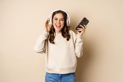 Happy caucasian woman listening music in headphones on smartphone app, dancing and having fun, standing over beige background.