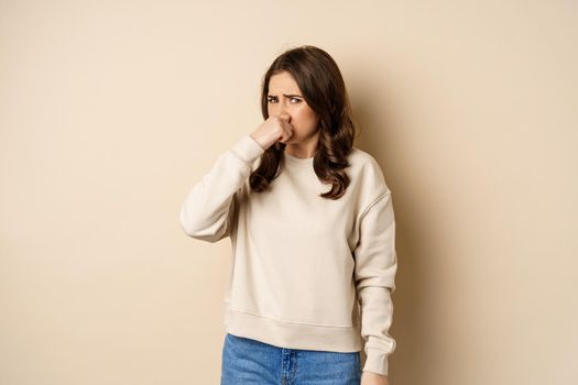 Woman expressing disgust of bad disgusting smell, close nose and grimaing from dislike, standing over beige background.