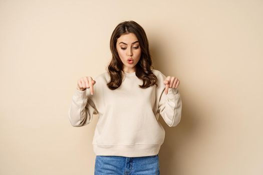 Surprised girl pointing and looking down at smth curious below, beige background.