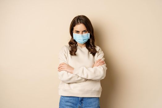Covid-19, pandemic and quarantine concept. Beautiful modern woman in medical face mask, cross arms on chest, looking confident, beige background.
