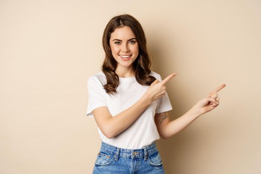 Smiling brunette woman in t-shirt, pointing fingers right, showing promo offer or advertisement, demonstrating banner, standing against beige background.