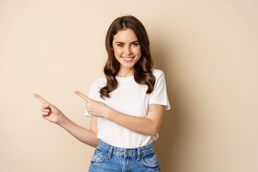 Stylish young caucasian woman smiling, pointing fingers left, showing advertisement, promo offer, standing against beige background.