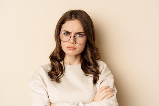 Close up portrait of complicated, troubled woman in glasses, frowning and looking displeased, standing over beige background. Copy space