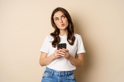 Portrait of woman thinking, holding mobile phone and looking up with thoughtful face, standing over beige background. Copy space