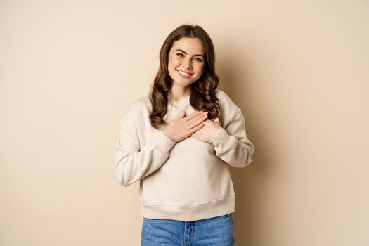 Smiling woman holding hands on heart, looking with care and love at camera, standing in sweater over beige background.