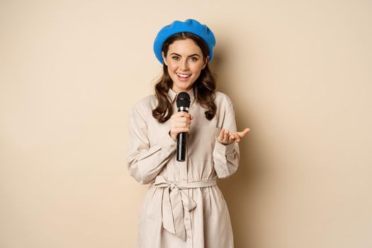 Portrait of happy stylish woman performing, singing with microphone, posing against beige background.