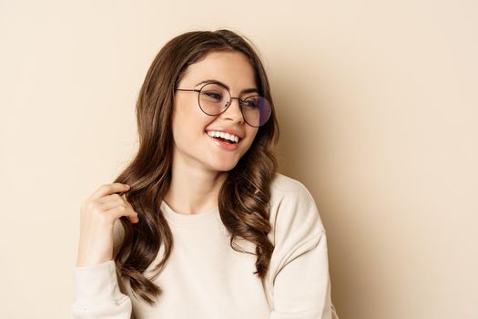 Close up portrait of stylish brunette woman in glasses, laughing and smiling, posing in eyewear against beige background.