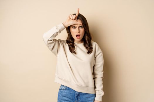 Woman showing loser sign on forehead, L word, standing over beige background. Copy space